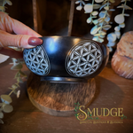 A hand with dark red nail polish holds a black ceramic bowl decorated with two white geometric flower-of-life patterns. The bowl is displayed on a wooden stand in a rustic setting with eucalyptus leaves and a shell in the background.