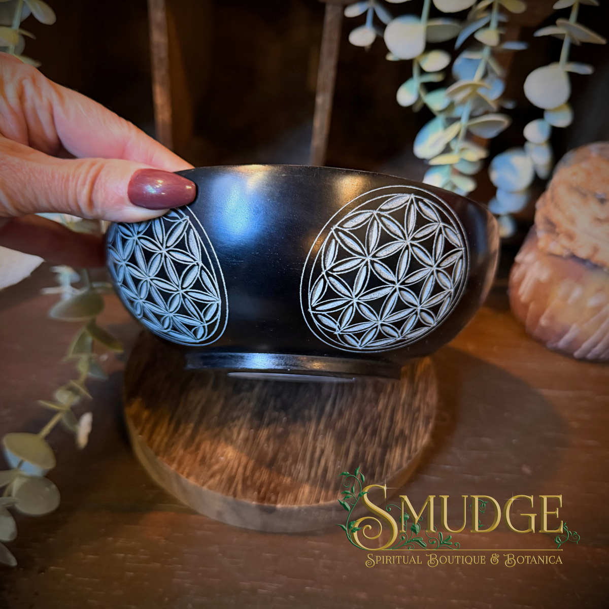 A hand with dark red nail polish holds a black ceramic bowl decorated with two white geometric flower-of-life patterns. The bowl is displayed on a wooden stand in a rustic setting with eucalyptus leaves and a shell in the background.