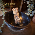 A smudge bundle wrapped in white string rests inside a dark ceramic bowl on a wooden surface, with decorative eucalyptus leaves and a warm rustic background. The brand name 'SMUDGE' is visible in the upper left corner.
