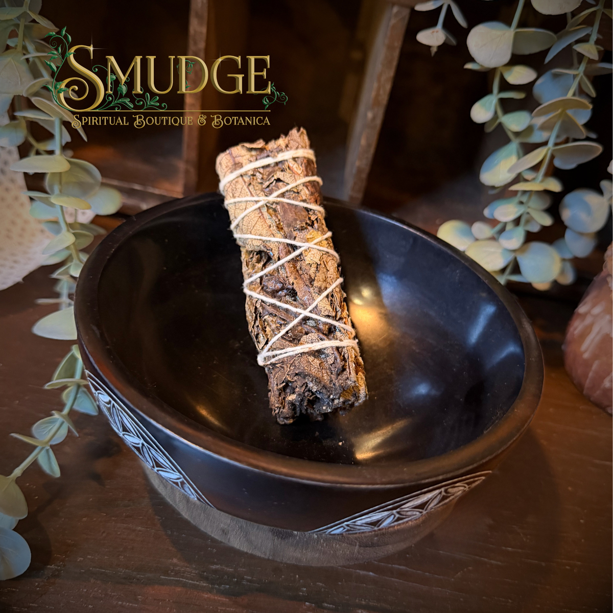 A smudge bundle wrapped in white string rests inside a dark ceramic bowl on a wooden surface, with decorative eucalyptus leaves and a warm rustic background. The brand name 'SMUDGE' is visible in the upper left corner.