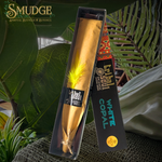 A packaged white copal incense stick from the brand Tribal Soul, displayed in its black and gold box next to the open incense holder. The incense stick is wrapped in gold paper with a bright yellow feather, set against a background of lush green tropical leaves.
