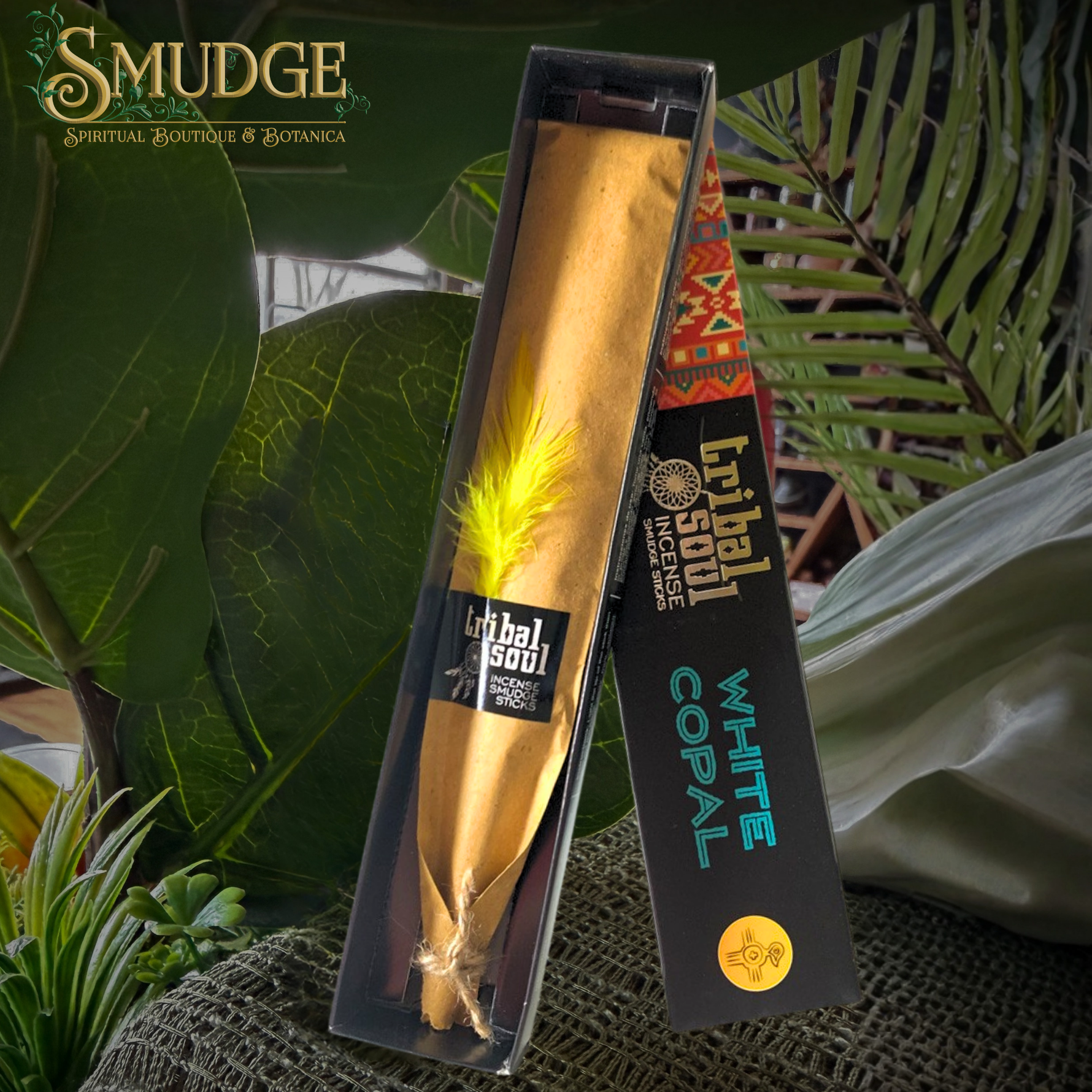 A packaged white copal incense stick from the brand Tribal Soul, displayed in its black and gold box next to the open incense holder. The incense stick is wrapped in gold paper with a bright yellow feather, set against a background of lush green tropical leaves.