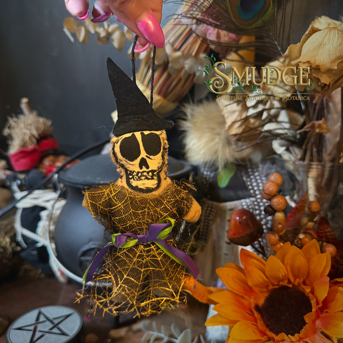A handmade Halloween hanging ornament shaped like a skeleton wearing a black witch hat and cobwebbed dress, held by a hand with pink manicured nails. The background is a dark, decorative boho setup with dried flowers, a cauldron, and a peacock feather, featuring a 'SMUDGE' brand watermark.