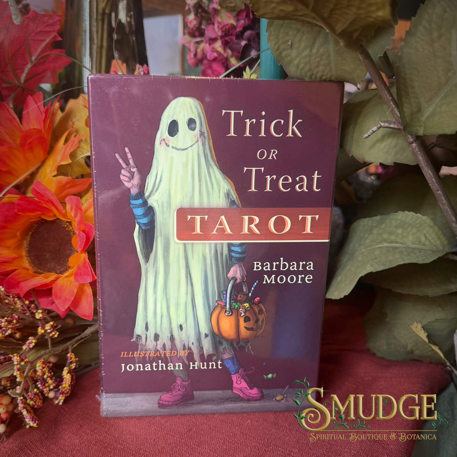 The book 'Trick or Treat Tarot' by Barbara Moore and Jonathan Hunt is displayed on a red fabric surface surrounded by autumn leaves and flowers. A 'Smudge Spiritual Marketplace & Botanica' logo is visible in the bottom right corner.