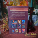 The back of a 'Trick or Treaters' card deck box is shown standing upright on a draped red tablecloth, surrounded by autumn-themed decorative props including a red glass vase, dried flowers, and a teal candle. The box is branded with the 'Smudge Spiritual Boutique & Botanica' logo in the bottom right corner.