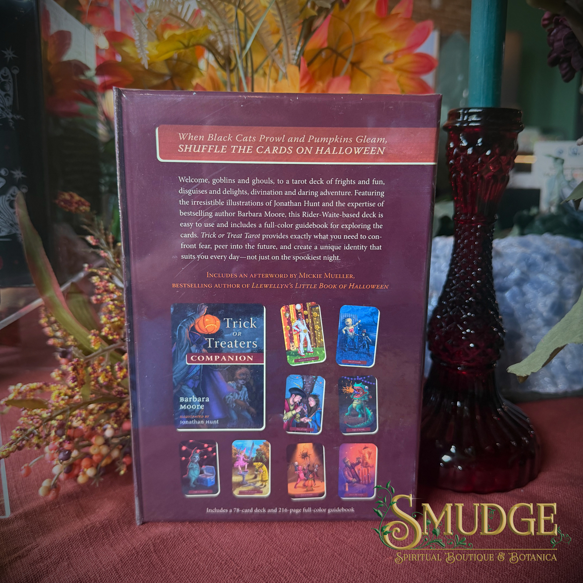 The back of a 'Trick or Treaters' card deck box is shown standing upright on a draped red tablecloth, surrounded by autumn-themed decorative props including a red glass vase, dried flowers, and a teal candle. The box is branded with the 'Smudge Spiritual Boutique & Botanica' logo in the bottom right corner.