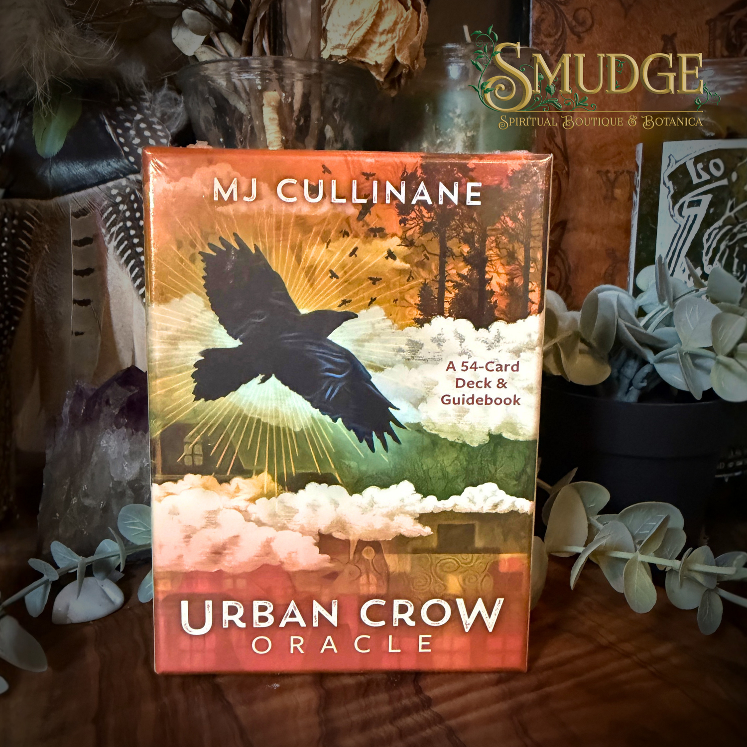 Urban Crow Oracle card deck box by MJ Cullinane, displayed on a wooden surface with dried botanicals, crystals, and greenery. The box features an illustration of a crow flying over clouds at sunset.