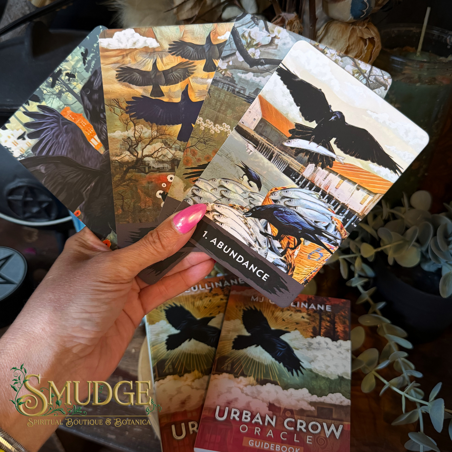 A hand with pink nail polish holds a fan of cards from the Urban Crow Oracle deck, showing illustrations of crows and a card titled 'Abundance.' The deck box and booklet are visible below, with decorative plants and a warm indoor setting in the background.