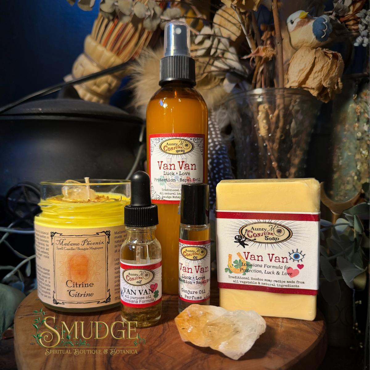 A collection of handmade natural products from Smudge Spiritual Shop arranged on a wooden board, including a yellow Citrine Glycerin candle, a brown amber spray bottle of Van Van Aromatic Spray, a small Van Van Aromatic Oil bottle, a Van Van Sacred Rose oil bottle, and a bar of Van Van Handmade Palm Soap.