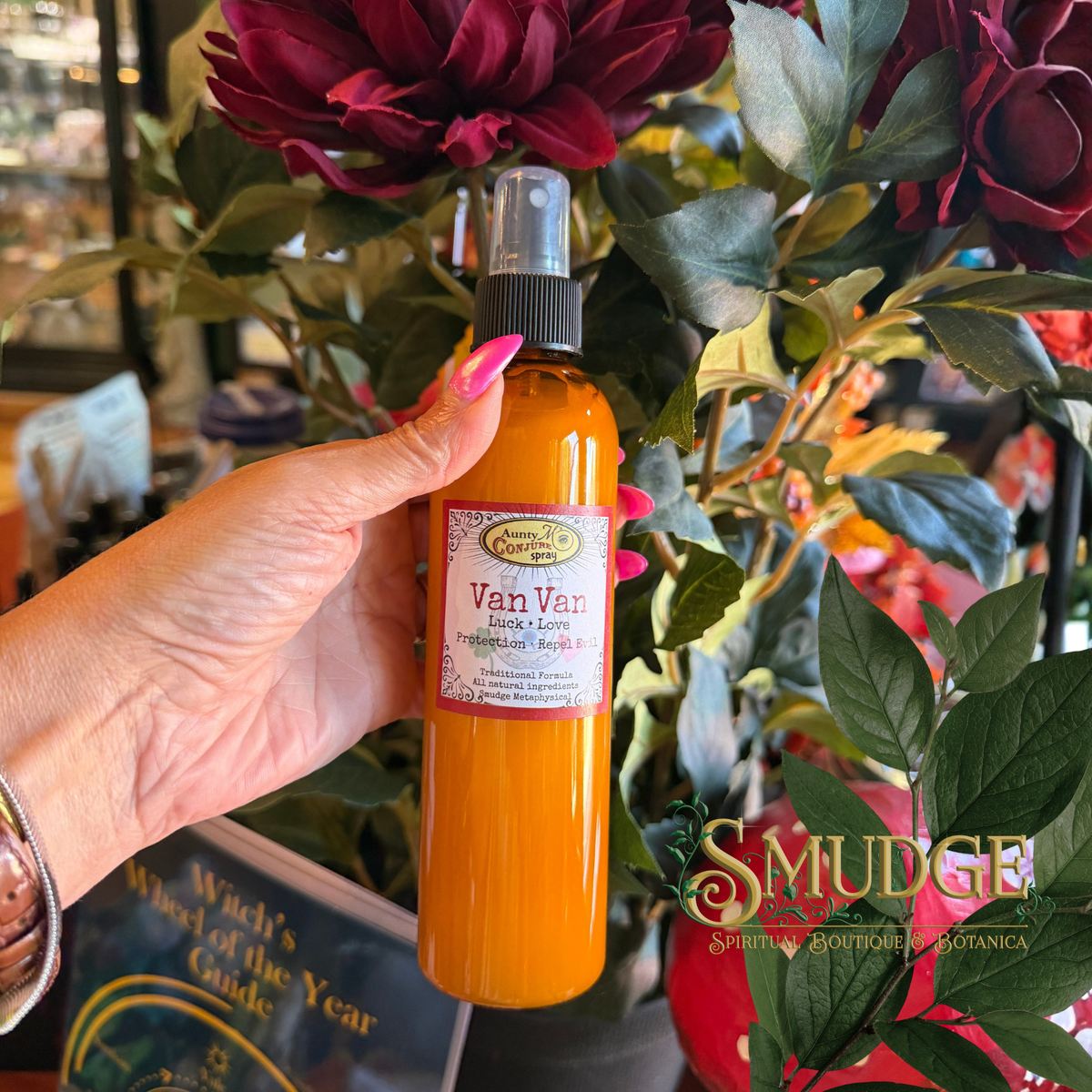 A hand with pink nails holds a bottle of Van Van Spiritual Balm by Smudge Spiritually Spiritual Botanicals in front of a background of red flowers and green leaves.