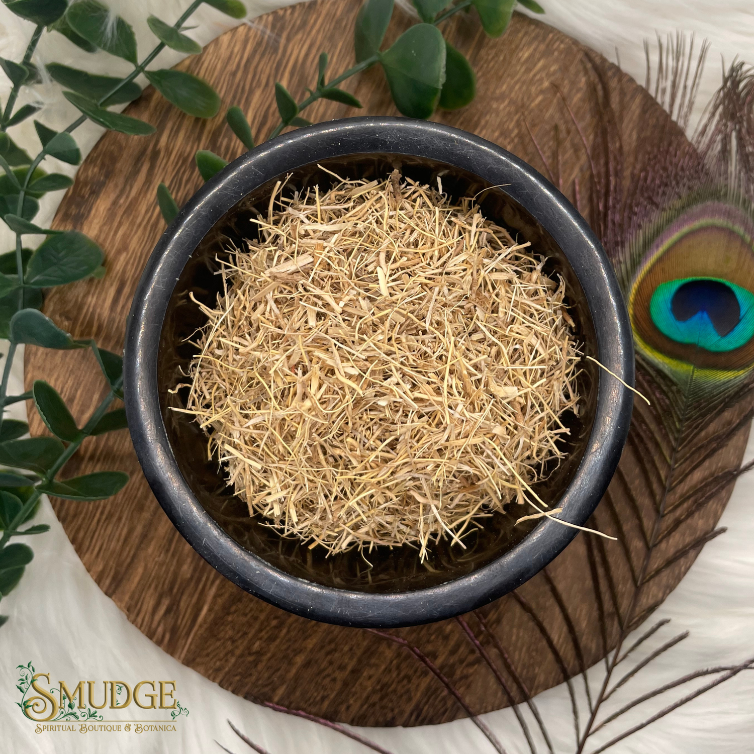 Black bowl with dried grass on a wooden surface, surrounded by greenery and a peacock feather.