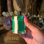 A hand with bright pink nails holds a small square glass bottle filled with bright green liquid. The bottle has a black cap and a white label reading 'Smudge Moisturizer.