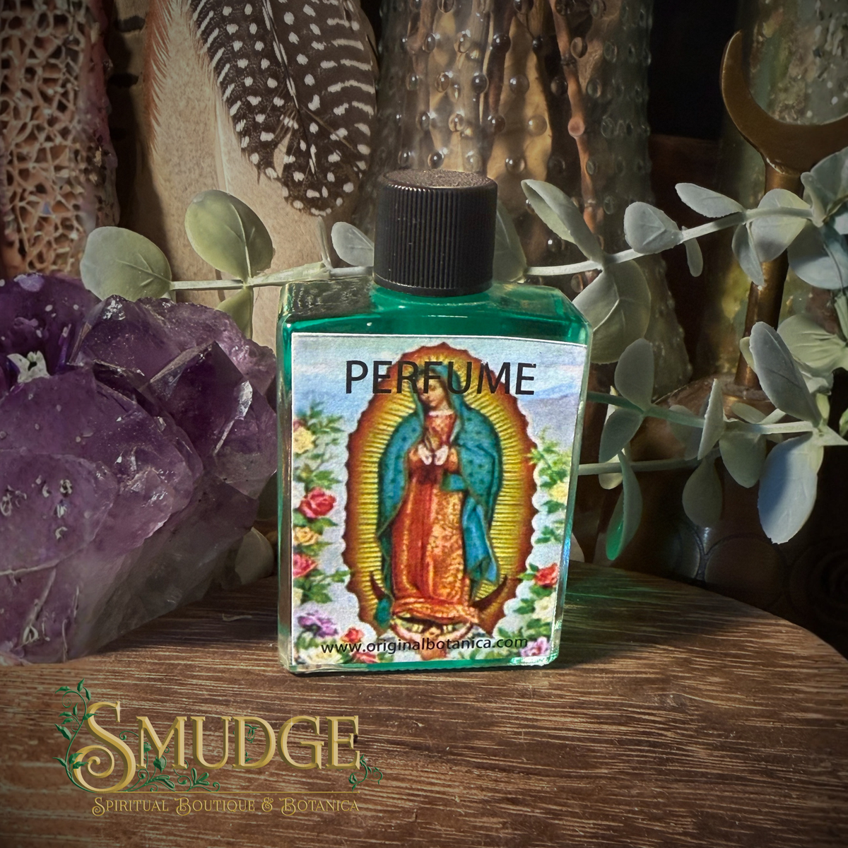 A small square glass bottle labeled 'PERFUME' with a colorful religious illustration of Our Lady of Guadalupe on the front, placed on a wooden surface with purple crystals, dried leaves, and textured decor in the background. The brand name 'Smudge' appears at the bottom left.