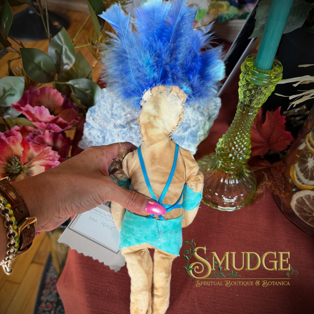 A handmade cloth doll with a tan body, wearing a turquoise dress and matching headpiece with bright blue feathers, held by a hand. The background features a green glass candle holder, flowers, and a red tablecloth with the brand logo 'Smudge Spiritual Boutique & Botanica'.
