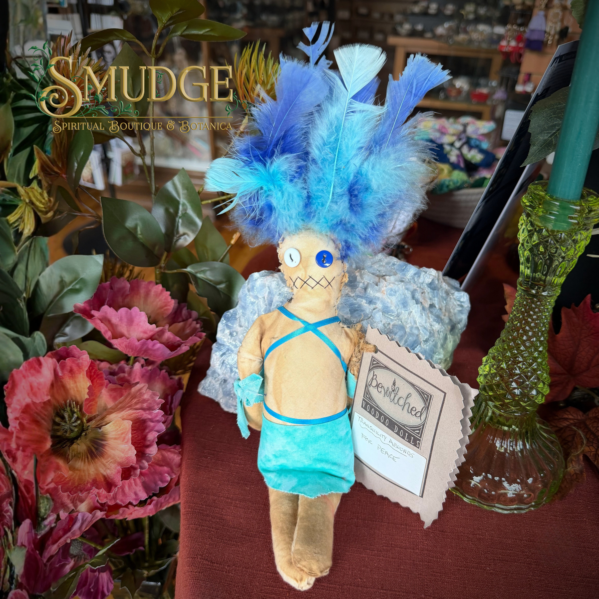 A handmade voodoo doll with a beige body, blue feather headdress, and turquoise clothing, displayed on a table with a 'Benwitch' tag and surrounded by flowers and decorative items.