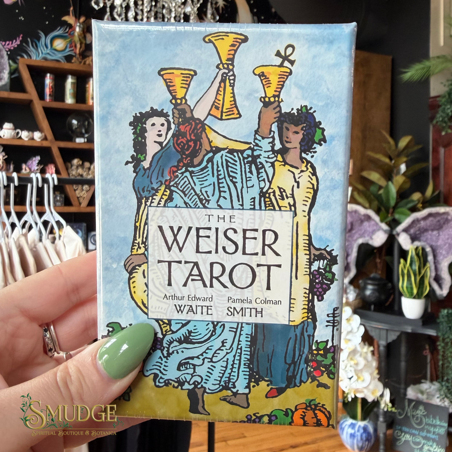 A hand with green nail polish holds a boxed tarot card deck in a retail shop setting. The box is labeled 'The Weiser Tarot' and features an illustrated cover with three figures holding gold cups against a light blue background.