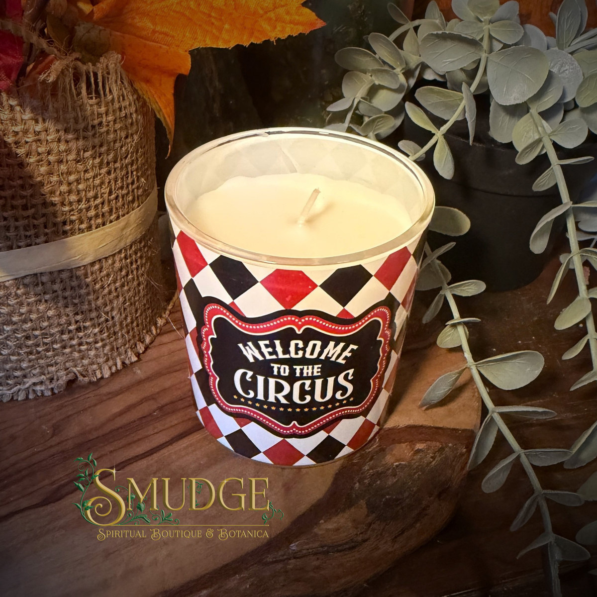 A white wax candle in a clear glass jar with a red, black, and white harlequin pattern label that reads 'Welcome to the Circus.' The candle is styled on a rustic wooden surface with burlap, greenery, and autumn leaves in the background.