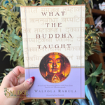 A hand holds a book titled 'What the Buddha Taught' by Walpola Rahula. The book cover has a cream-colored top section with Sanskrit text, a gold-toned Buddha face illustration in the center, and a lavender-purple bottom section.