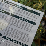 A close-up of a printed guidebook page titled 'Spiritual Application for Beginners' with sections on 'Altar' and 'Spellingcast,' photographed outdoors against a blurred green and gold background. The Mudge brand logo is visible in the top right corner.