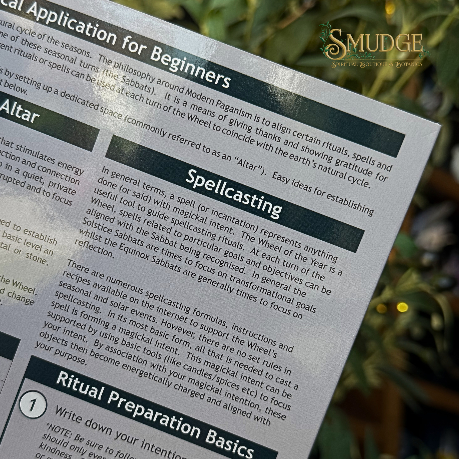 A close-up of a printed guidebook page titled 'Spiritual Application for Beginners' with sections on 'Altar' and 'Spellingcast,' photographed outdoors against a blurred green and gold background. The Mudge brand logo is visible in the top right corner.