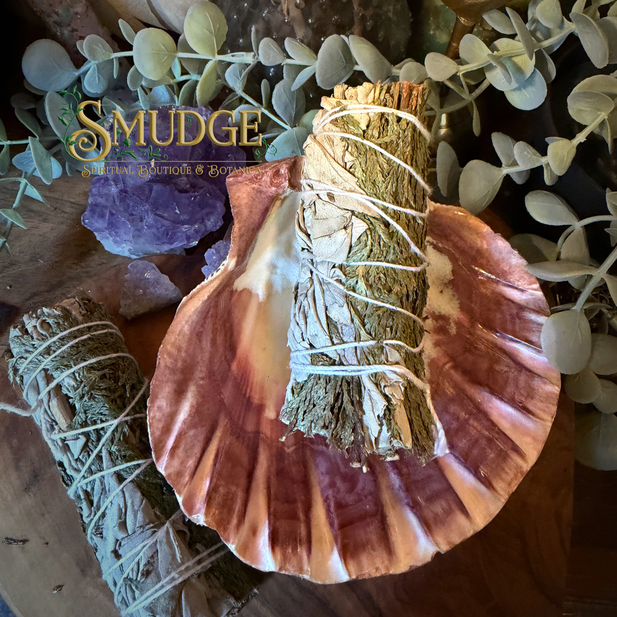 A styled product photo showing a large seashell with a cedar and white sage smudge bundle placed inside, another sage bundle on the left, crystals, eucalyptus leaves, and a wooden surface. The brand name 'SMUDGE' is visible as an overlay.