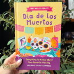 A hand holds a book titled 'Why We Celebrate Día de los Muertos' against a background of green foliage. The book cover features a bright yellow top half with purple title text, a pink banner, and colorful illustrations of sugar skulls, candles, and papel picado.