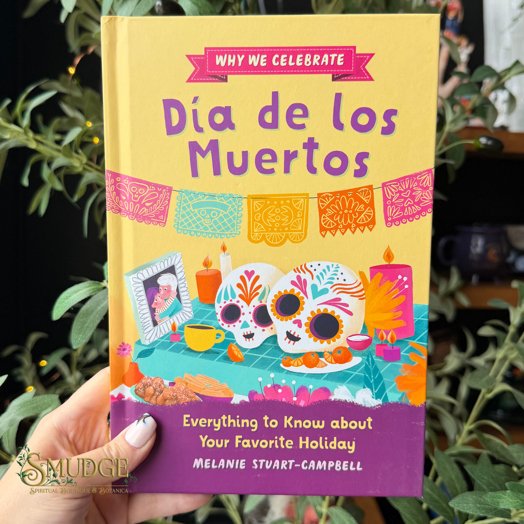 A hand holds a book titled 'Why We Celebrate Día de los Muertos' against a background of green foliage. The book cover features a bright yellow top half with purple title text, a pink banner, and colorful illustrations of sugar skulls, candles, and papel picado.