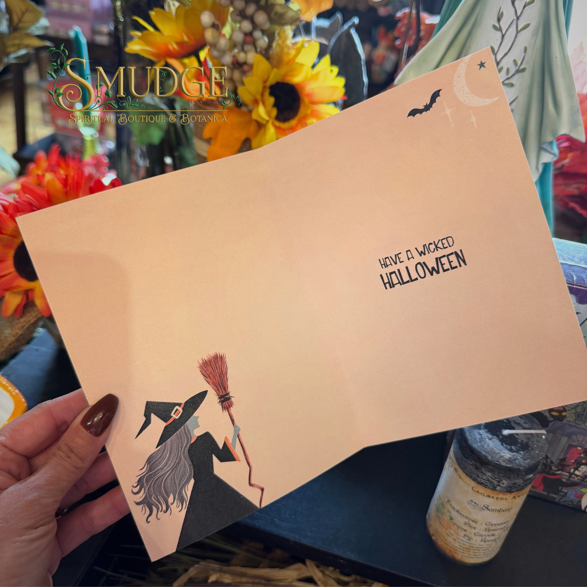 A hand holds a peach-colored greeting card featuring a witch illustration and the text 'HAVE A WICKED HALLOWEEN' on a decorated craft display background.