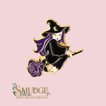 A witch-themed enamel pin featuring a black-robed witch with purple hair riding a broom, set against a light pink background. The pin has gold-tone metal outlines and is displayed above the 'SMUDGE Spiritual Boutique & Botanica' logo.