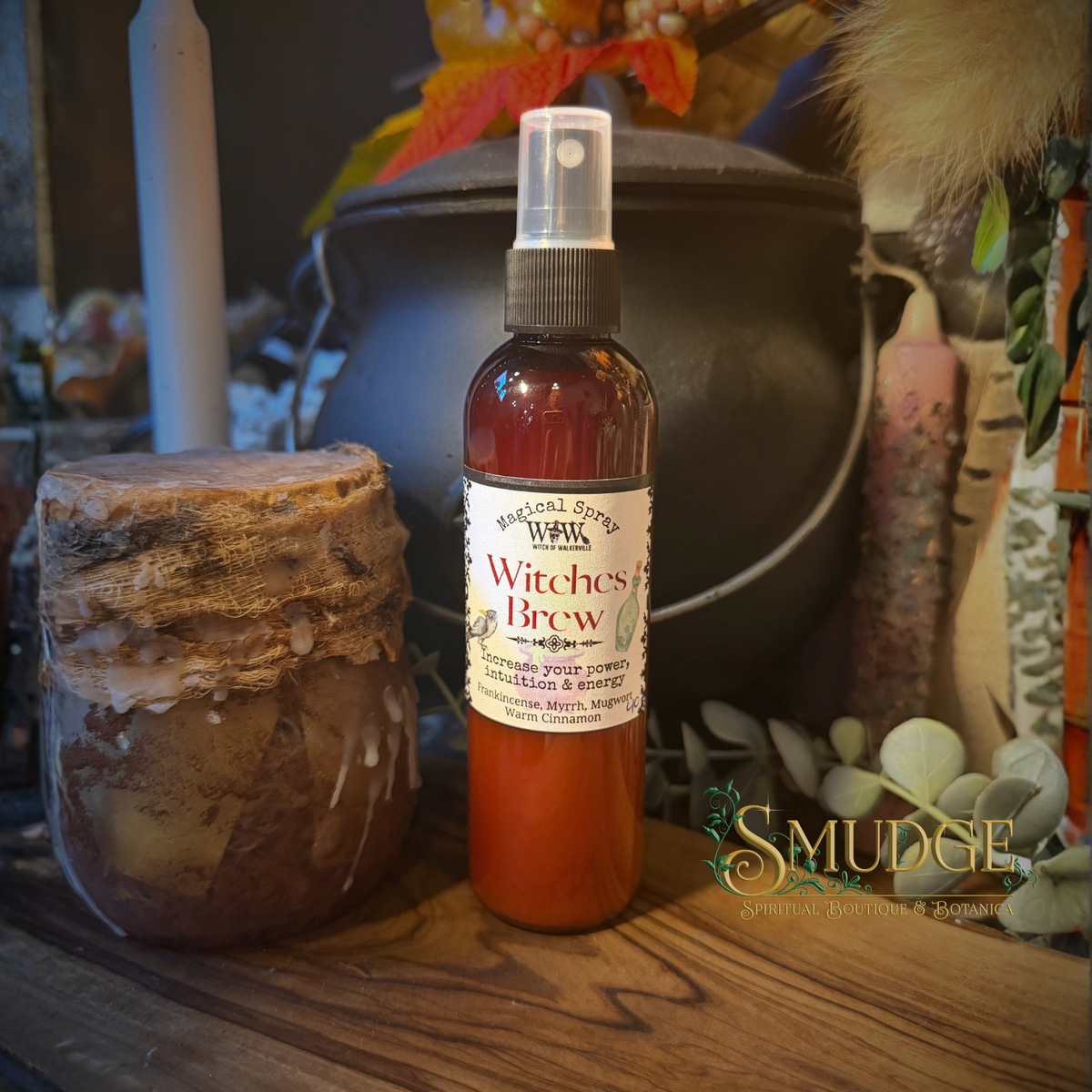 A bottle of Witches Brew essential oil spray with amber liquid and a white label, styled on a wooden surface next to a jar with a cork lid in a rustic autumn-themed setting.