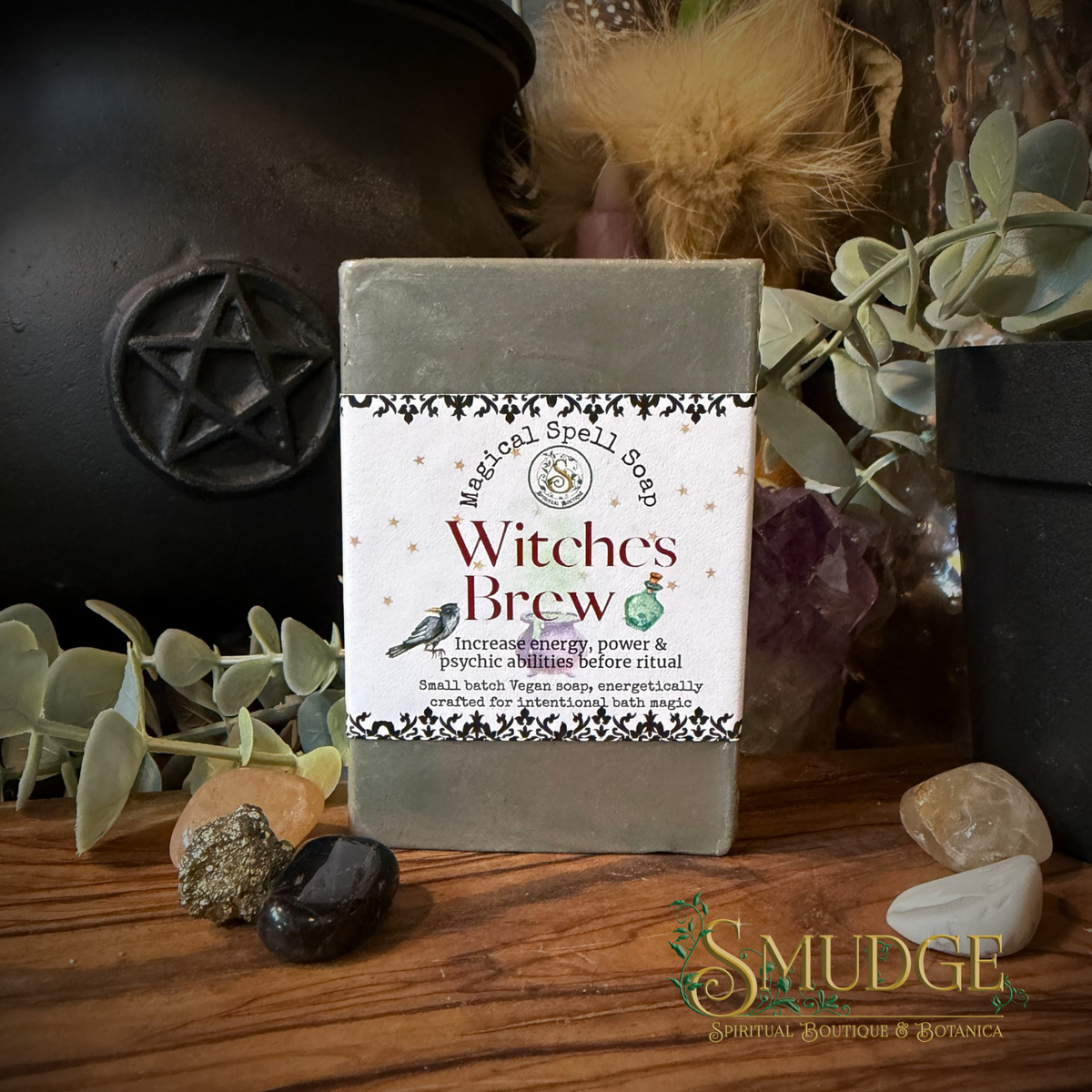 A bar of witches brew soap on a wooden table with a black cauldron and eucalyptus behind it. 
