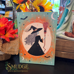 A rectangular decorative card or print featuring a witch in a black dress and hat holding a broom, surrounded by orange circular linework, stars, bats, and crescent moons on a pale aqua background. The card is photographed on a tabletop with flowers and store decor around it.