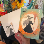 A hand holds a printed card featuring a witch silhouette with a broom inside an orange circular frame, with a second card behind it showing a matching witch illustration. The scene includes flowers and decorative objects in the background, and a visible brand watermark reads Smudge Southern Botanica.