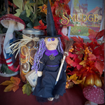 A witch-themed rag doll with a black hat and dress, purple yarn hair, and a broom, displayed in an autumn-themed decorative setting with fall leaves, mushrooms, and a jar of dried citrus slices.