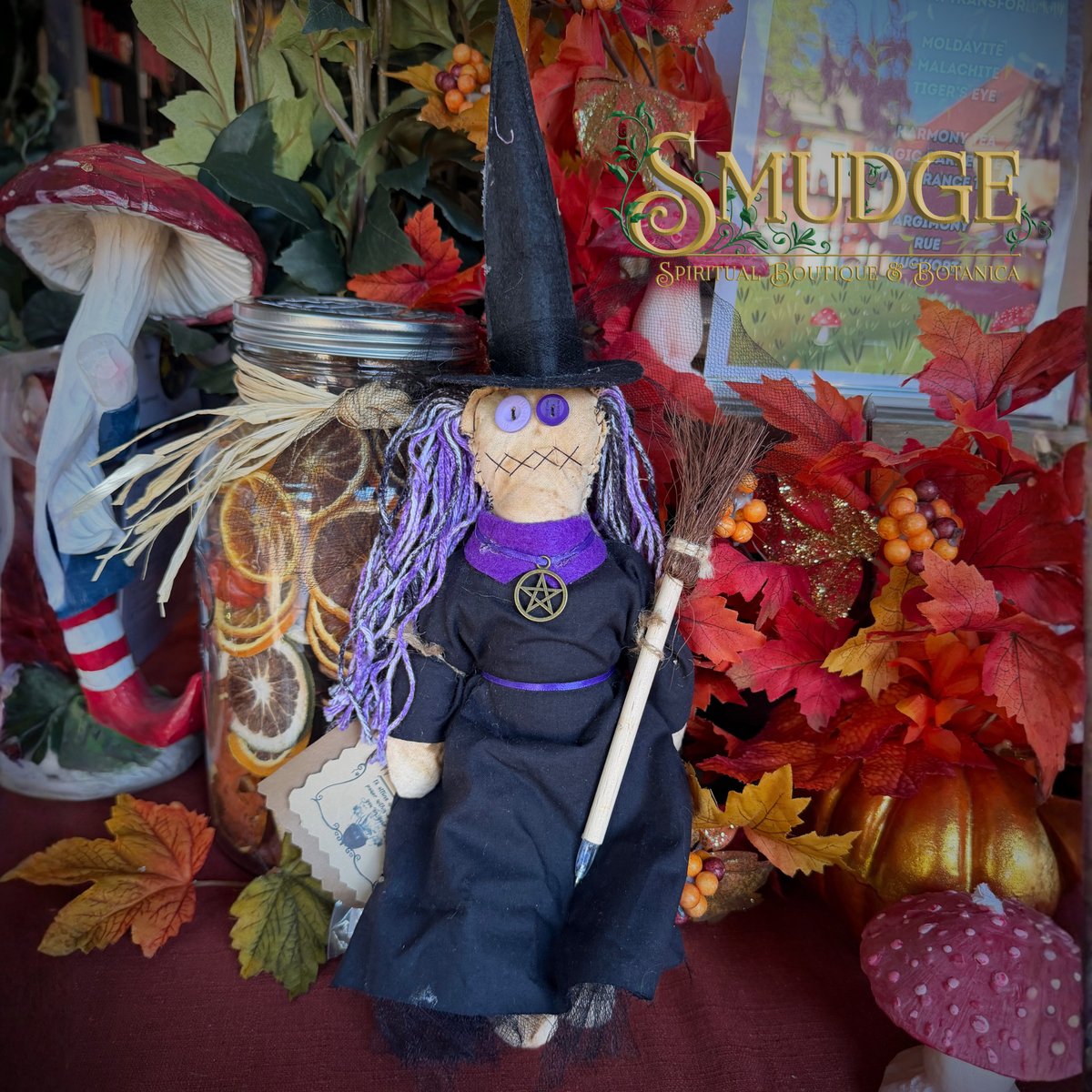A witch-themed rag doll with a black hat and dress, purple yarn hair, and a broom, displayed in an autumn-themed decorative setting with fall leaves, mushrooms, and a jar of dried citrus slices.
