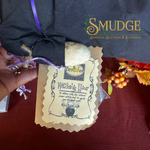 A hand holds a kraft paper tag attached to a black cloth pouch with a purple ribbon, set on a dark red surface. The tag reads 'Bewitched Prop Shop' and 'Watching Your'.