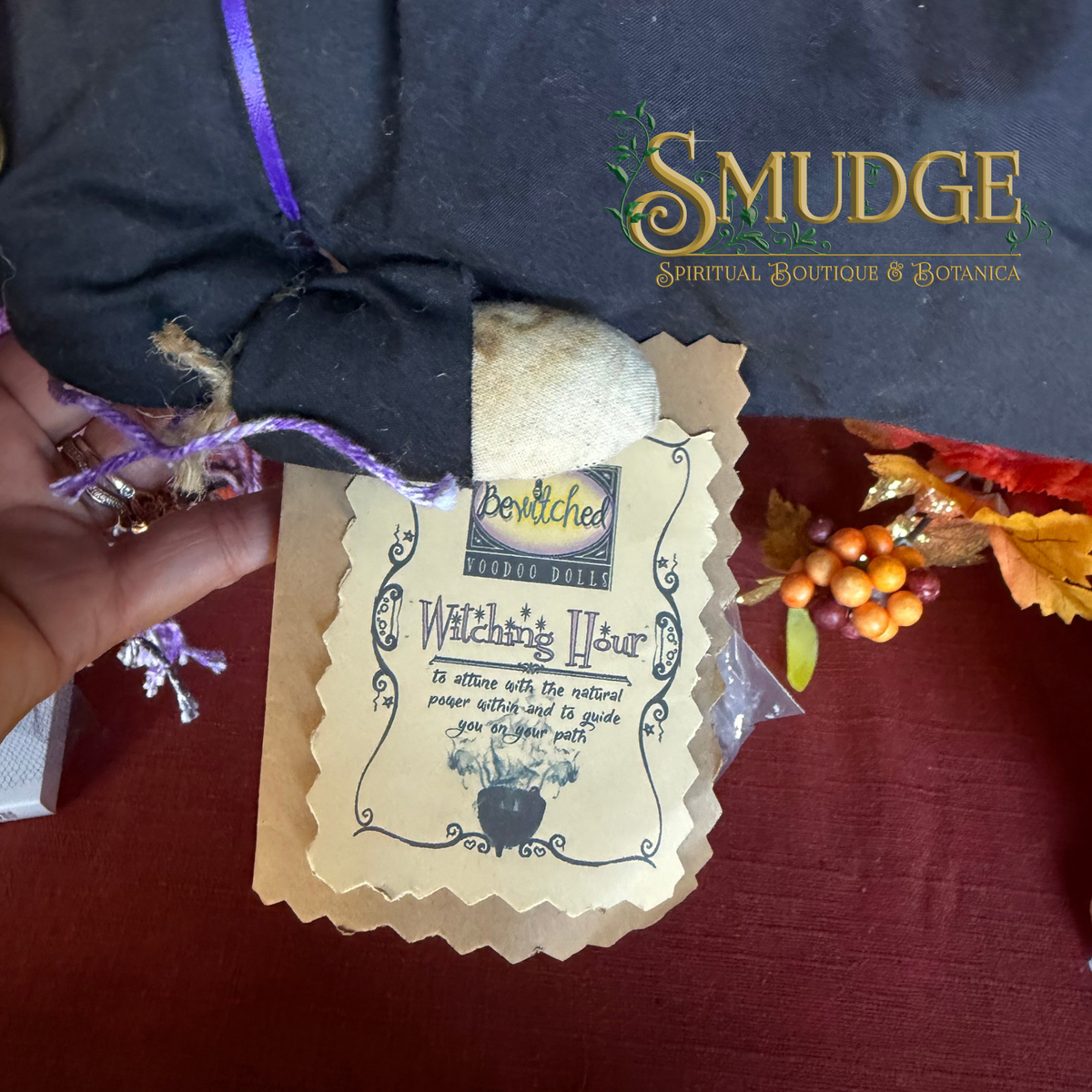 A hand holds a kraft paper tag attached to a black cloth pouch with a purple ribbon, set on a dark red surface. The tag reads 'Bewitched Prop Shop' and 'Watching Your'.