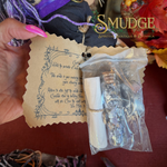 A hand with pink nails holds a small clear plastic bag containing dried botanical pieces and a white scroll, next to a tan paper card with printed text. A large 'Smudge Spiritual Boutique & Botanica' logo is visible in the upper right, and autumn leaves and purple fabric appear in the background.