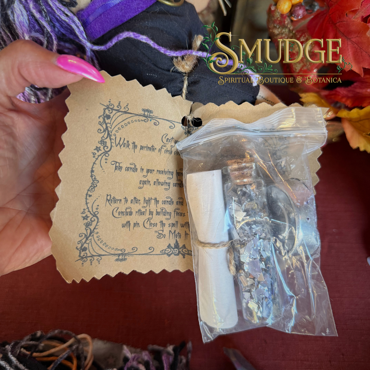 A hand with pink nails holds a small clear plastic bag containing dried botanical pieces and a white scroll, next to a tan paper card with printed text. A large 'Smudge Spiritual Boutique & Botanica' logo is visible in the upper right, and autumn leaves and purple fabric appear in the background.
