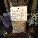 A clear plastic bag filled with dried yellow-brown herbs sits on a dark tabletop, wrapped with a white paper label reading 'Magical Herbs' and 'Yarrow'. The scene is styled with greenery, crystals, and decorative metal objects in a moody indoor setting.