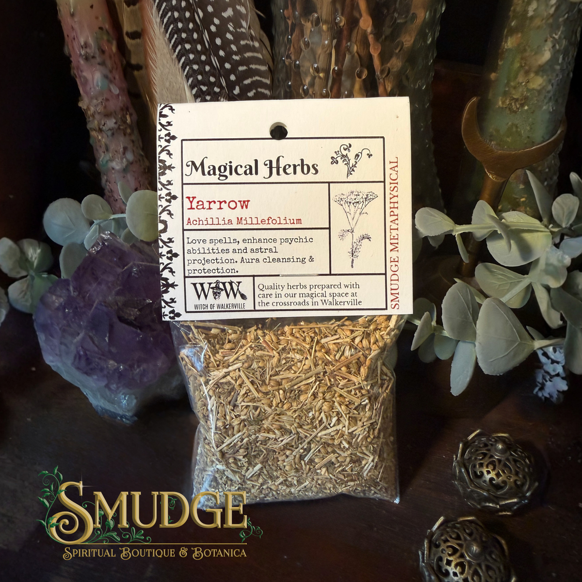 A clear plastic bag filled with dried yellow-brown herbs sits on a dark tabletop, wrapped with a white paper label reading 'Magical Herbs' and 'Yarrow'. The scene is styled with greenery, crystals, and decorative metal objects in a moody indoor setting.