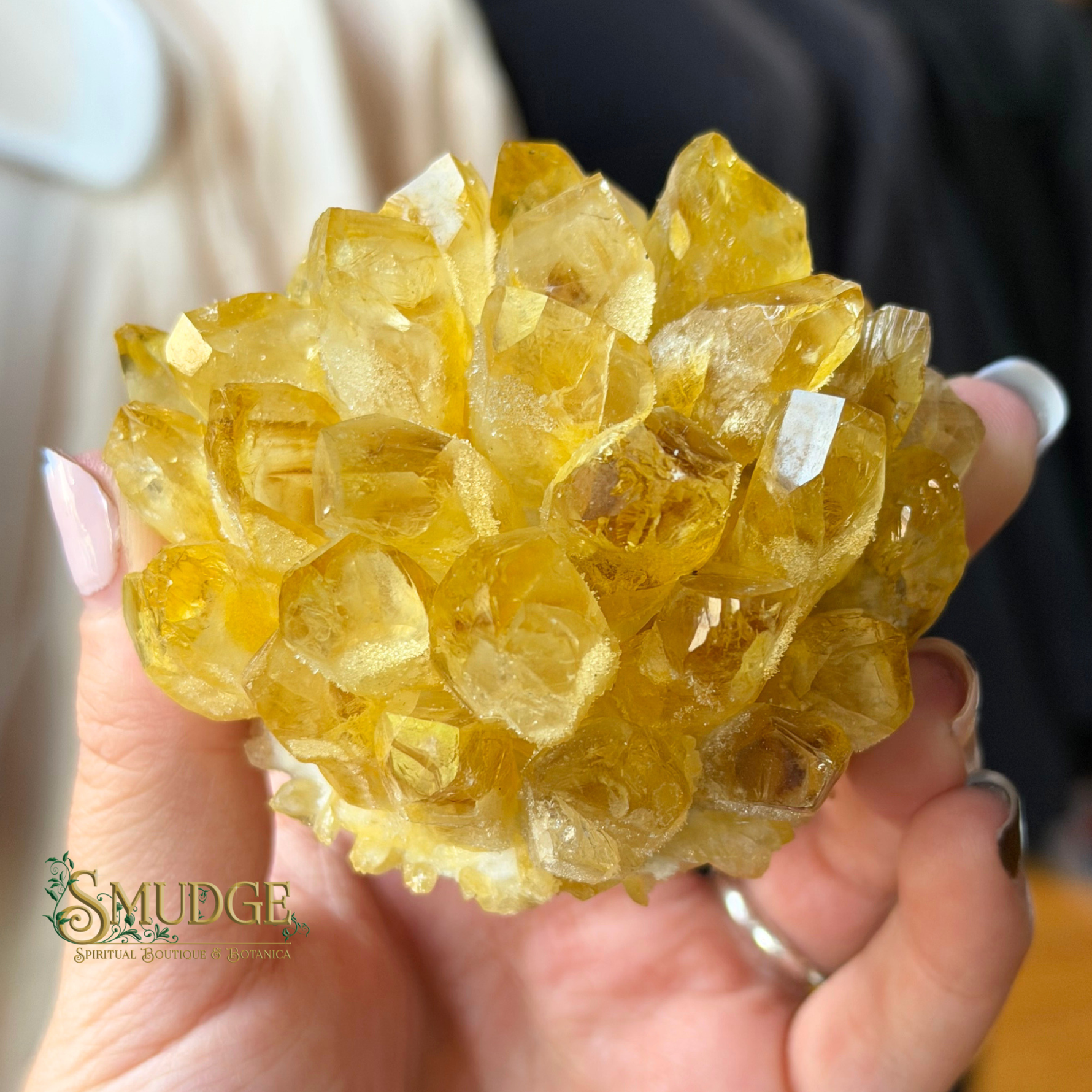 A hand holding a cluster of translucent yellow citrine crystals, with a 'SMUDGE Gemstone Collection' logo watermark in the lower-left corner.