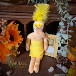 A handmade voodoo doll with a yellow feathered wig, yellow dress, and yellow buttons for eyes, displayed on a wooden surface with autumn and botanical props. The brand name 'SMUDGE Spiritual Boutique & Botany' is visible in the bottom left corner.