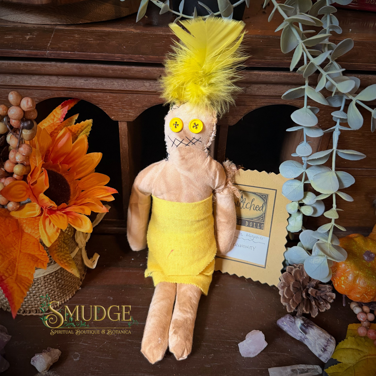 A handmade voodoo doll with a yellow feathered wig, yellow dress, and yellow buttons for eyes, displayed on a wooden surface with autumn and botanical props. The brand name 'SMUDGE Spiritual Boutique & Botany' is visible in the bottom left corner.