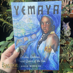 Book titled 'Yemaya: Orisha, Goddess, and Queen of the Sea' held by a hand with green nail polish against a plant background.