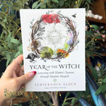 A hand holds a book titled 'Year of the Witch' by Temperance Alden. The book cover features a white background with a detailed illustration of a circular wreath made of branches, surrounding a sun face and various colorful seasonal elements.